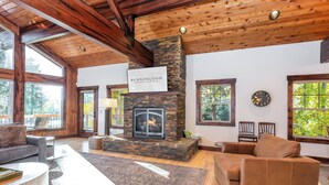 A spacious living room with a stone fireplace, wooden ceiling beams, and large windows offering views of the outdoors. - PONY47 - Stunning River Front Home, Community Pool (Sunriver)