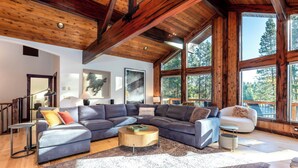 A spacious living room with a large sectional sofa, a round coffee table, and a view of the outdoors through large windows. - PONY47 - Stunning River Front Home, Community Pool (Sunriver)