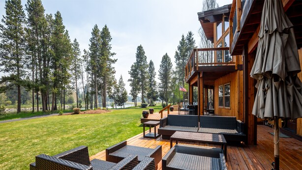 A wooden deck with wicker furniture, overlooking a grassy area and tall trees. - PONY47 - Stunning River Front Home, Community Pool (Sunriver)
