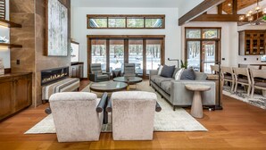 A spacious living room with a fireplace, comfortable seating, and large windows offering a view of the outdoors. - SUNS93 - Gorgeous Caldera Springs Home (Sunriver)