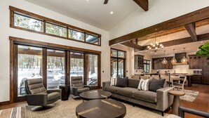 A spacious living area with a large sofa, two armchairs, and a coffee table. There are large windows offering a view of a snowy landscape. The room has wooden beams on the ceiling and a modern kitchen in the background. - SUNS93 - Gorgeous Caldera Springs Home (Sunriver)