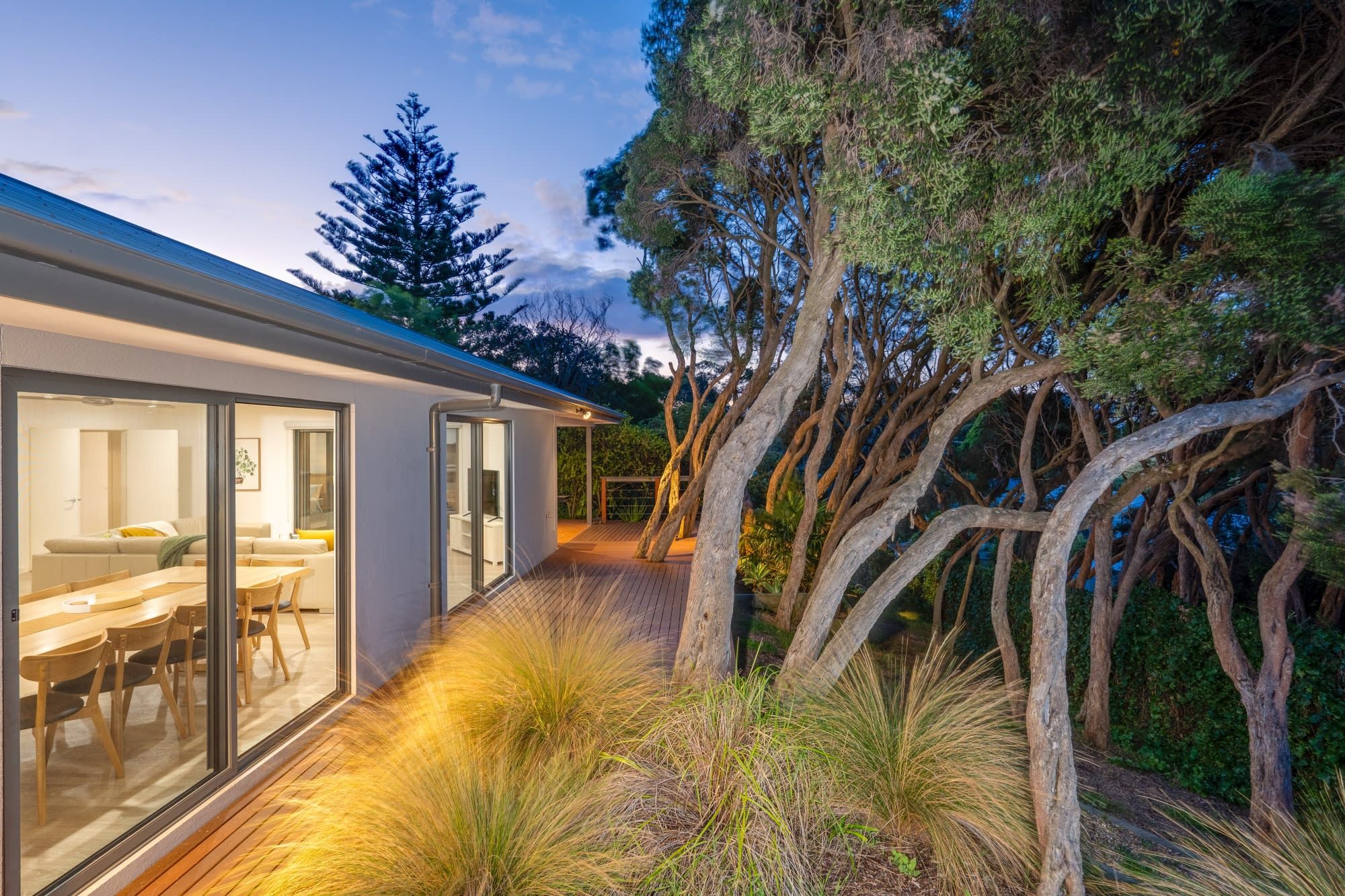A modern house with a glass-enclosed dining area, a wooden deck, and a garden with trees and grasses.