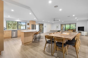 A modern kitchen with a central island, a dining area with a large table and chairs, and a living space with a sofa and TV.