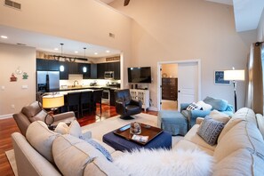 A modern living room with a sofa, a coffee table, and a TV. There is a kitchen area with a microwave and a refrigerator.