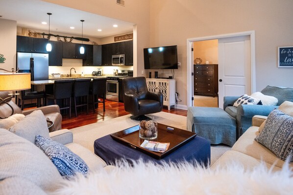 A modern living room with a sofa, a coffee table, and a TV. There is a kitchen area with dark cabinets and a microwave.
