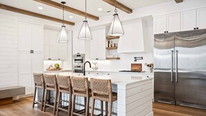 A modern kitchen with white cabinetry, a stainless steel refrigerator, and a central island with bar stools. - Vinnie's Beach House: Private Pool, Lagoon + Beaches (St George)