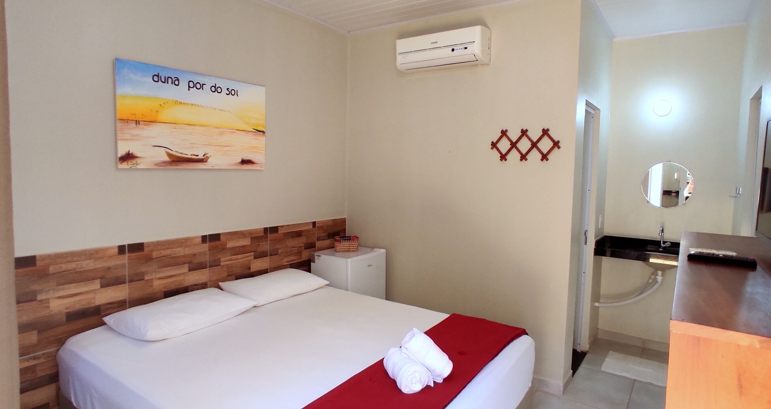 Una camera d'albergo con un letto, un comodino, un condizionatore d'aria a muro, un bagno con lavandino e specchio, e un elemento decorativo sulla parete.