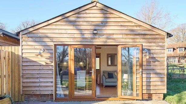 A wooden cabin with a sliding glass door entrance, a small porch, and a visible living area with a sofa and a lamp. - Host & Stay | Cobnut Barn (Bonnington)