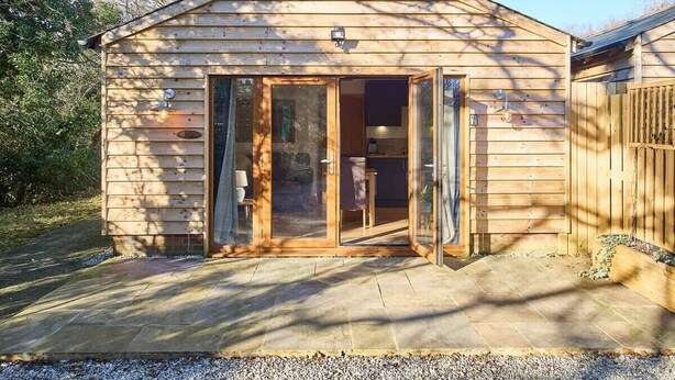 A wooden cabin with a glass door entrance, a small porch, and a stone patio. - Host & Stay | Oak Barn (Bonnington)