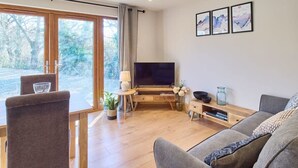 A modern living room with a large TV, grey sofa, and a dining area with a table and chairs. - Host & Stay | Oak Barn (Bonnington)