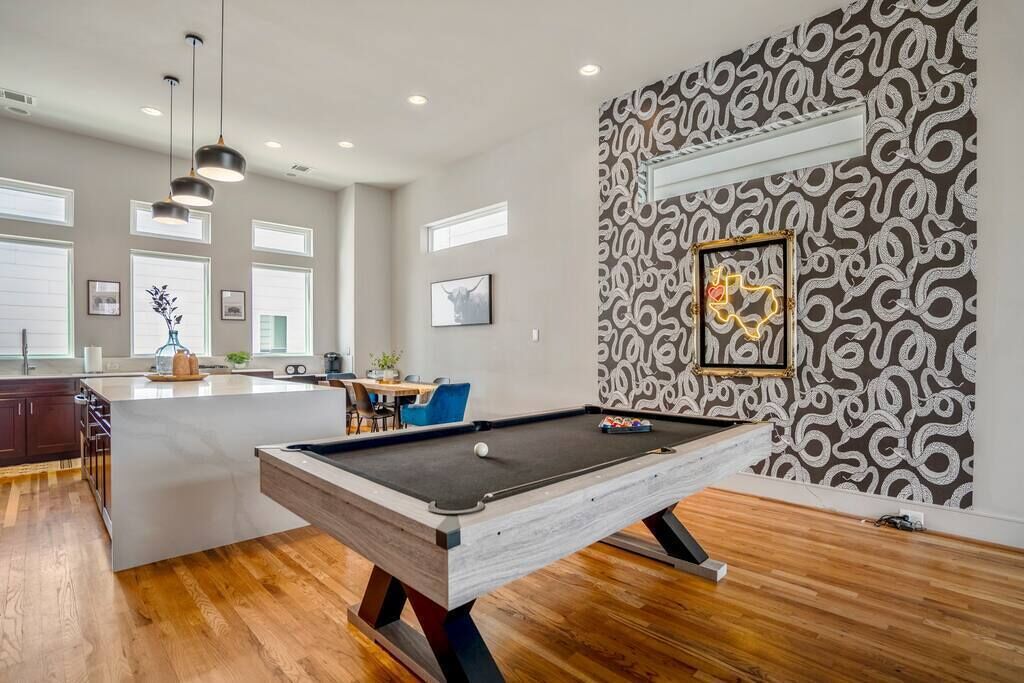 A pool table in a spacious living area with a modern design, a kitchen with dark wood cabinets, and a patterned wallpaper on the adjacent wall.