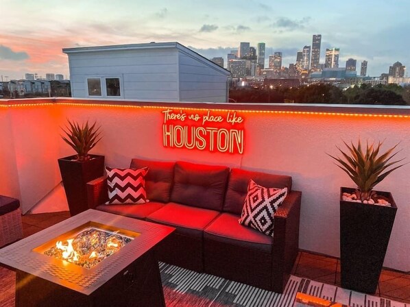 A rooftop lounge with a neon sign reading "There's no place like HOUSTON," a red sofa with patterned cushions, two potted plants, and a fire feature. - Luxury Downtown Getaway With Breathtaking Views (Houston)