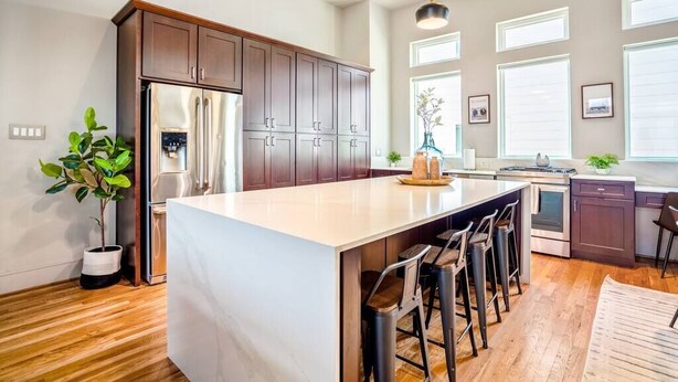 A modern kitchen with dark wooden cabinets, a large island with bar stools, stainless steel appliances, and large windows providing natural light. - Luxury Downtown Getaway With Breathtaking Views (Houston)