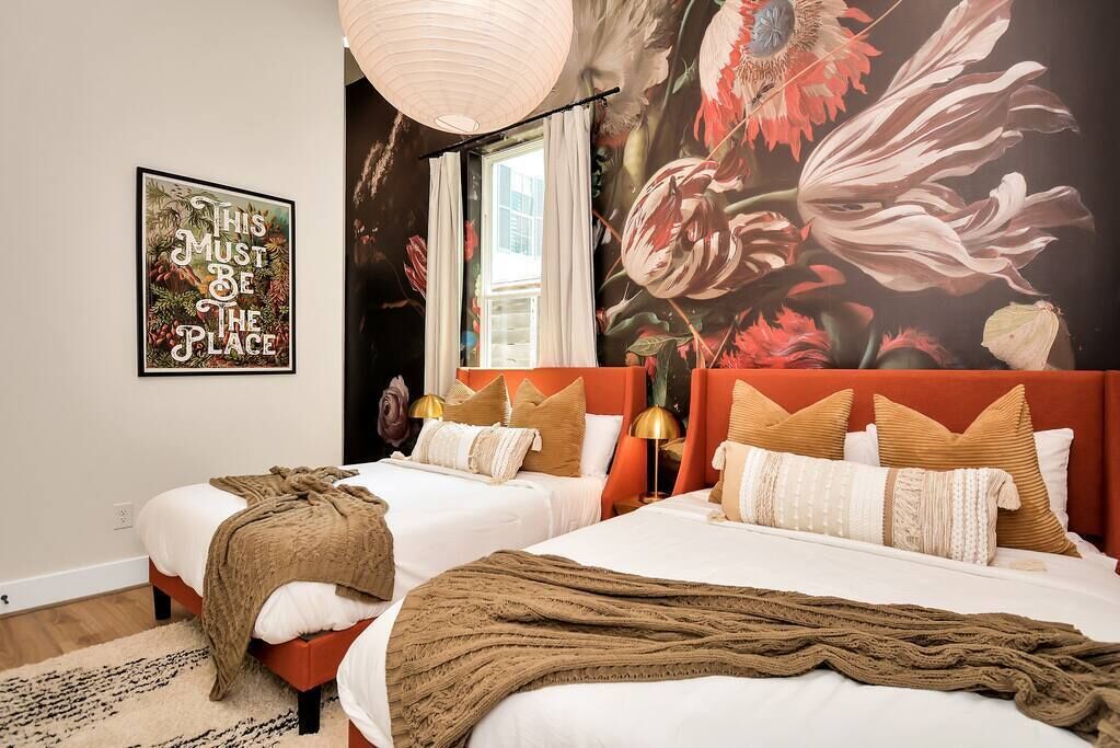 A bedroom with two beds, a floral mural, a framed quote on the wall, and a window with curtains.