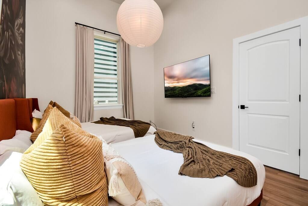 A hotel room with two beds, a large window with blinds, a painting on the wall, and a white door.