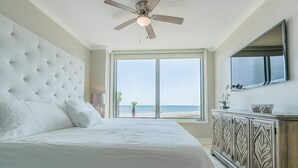 Ein Schlafzimmer mit einem großen Bett, einem Deckenventilator, einem großen Fenster mit Blick auf den Strand und einem Holzkasten.