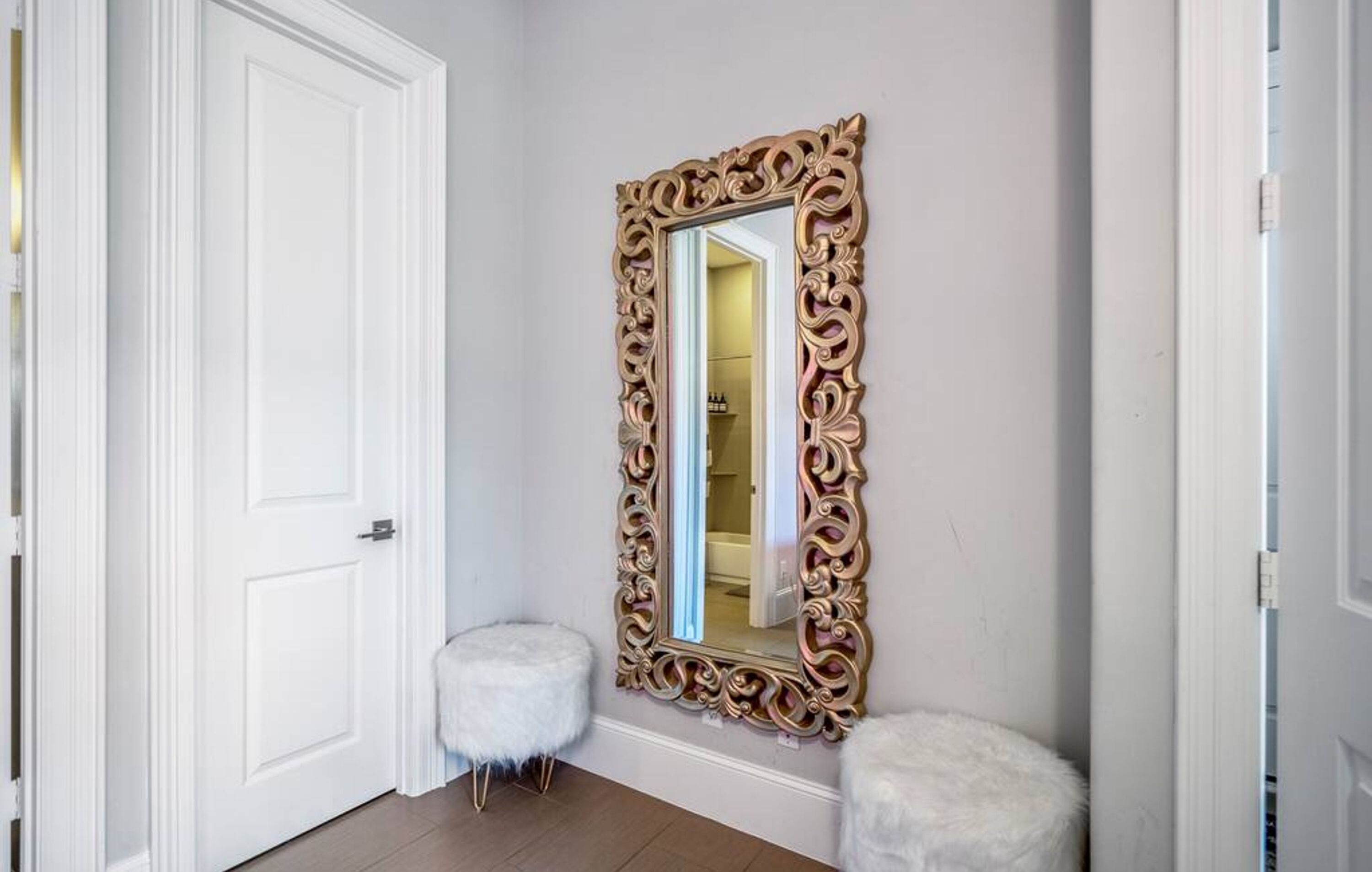 A hallway with a white door, a large ornate mirror, and a small white stool.