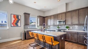 A modern kitchen with dark wood cabinets, a stainless steel island, and a bar seating area with orange chairs. - Luxury Downtown Home w Rooftop Deck in the Skyline (Houston)
