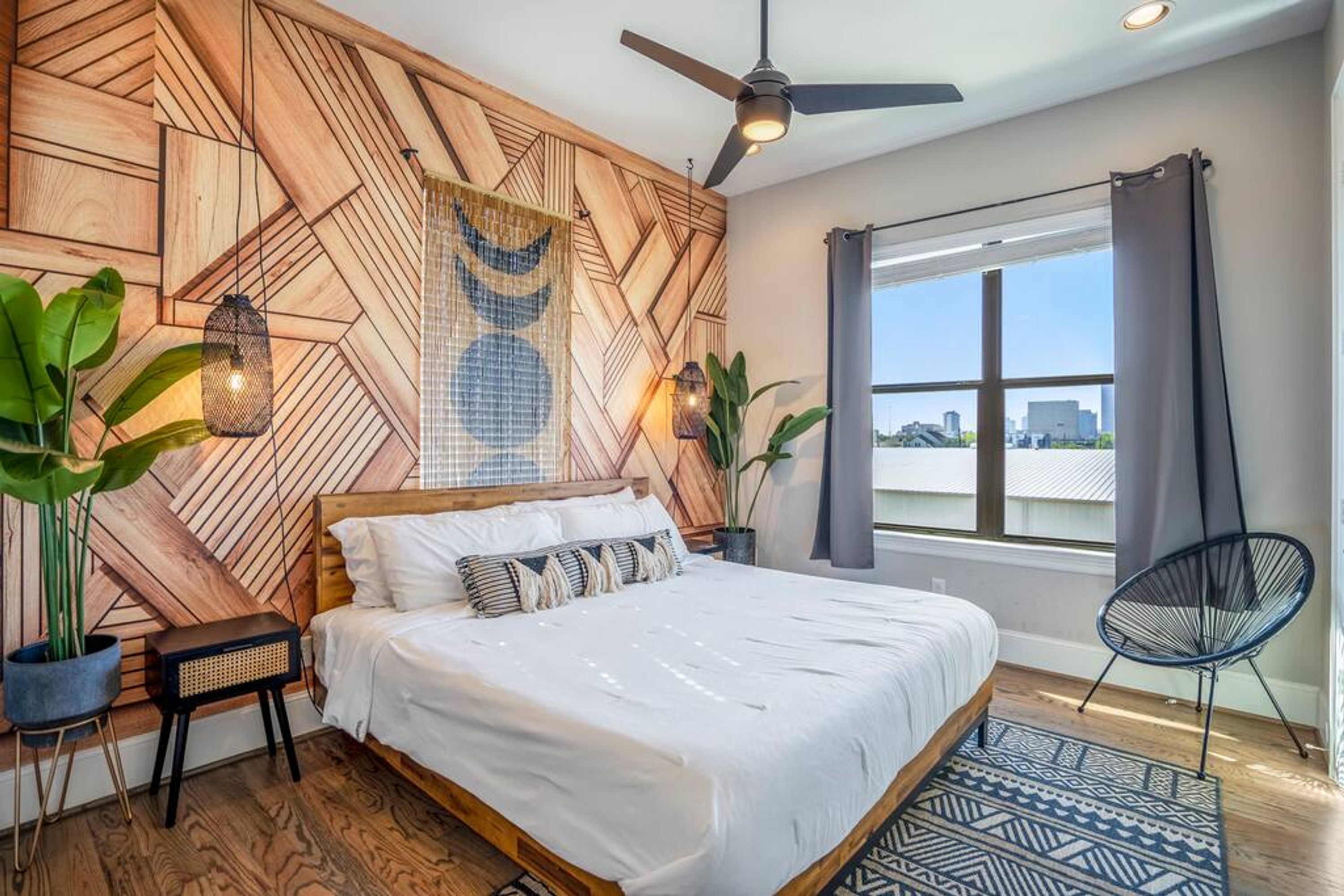 Une chambre moderne avec un mur en bois aux motifs géométriques, un lit avec une literie blanche, un ventilateur de plafond et une vue sur la ville par une grande fenêtre.