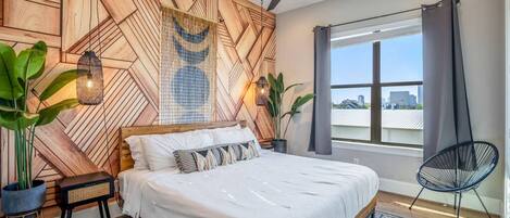 Une chambre moderne avec un mur en bois aux motifs géométriques, un lit avec une literie blanche, un ventilateur de plafond et une vue sur la ville par une grande fenêtre.