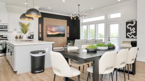 A modern kitchen with white cabinetry, a central island, and a dining area with a dark table and white chairs. - Paradise Palace Best Rooftop in HTX w Hot Tub (Houston)