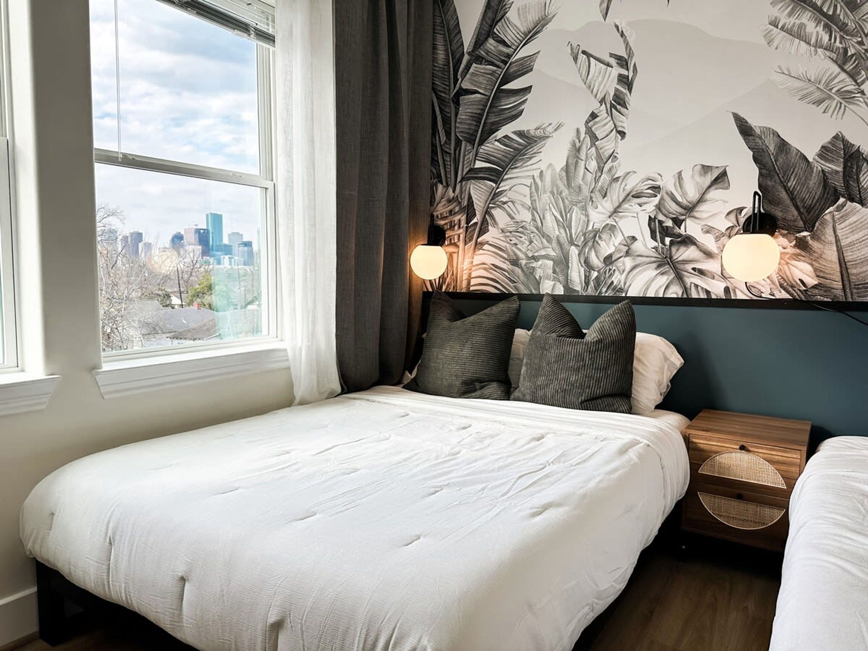 A bedroom with a city view, a large plant mural, and a nightstand with a lamp.