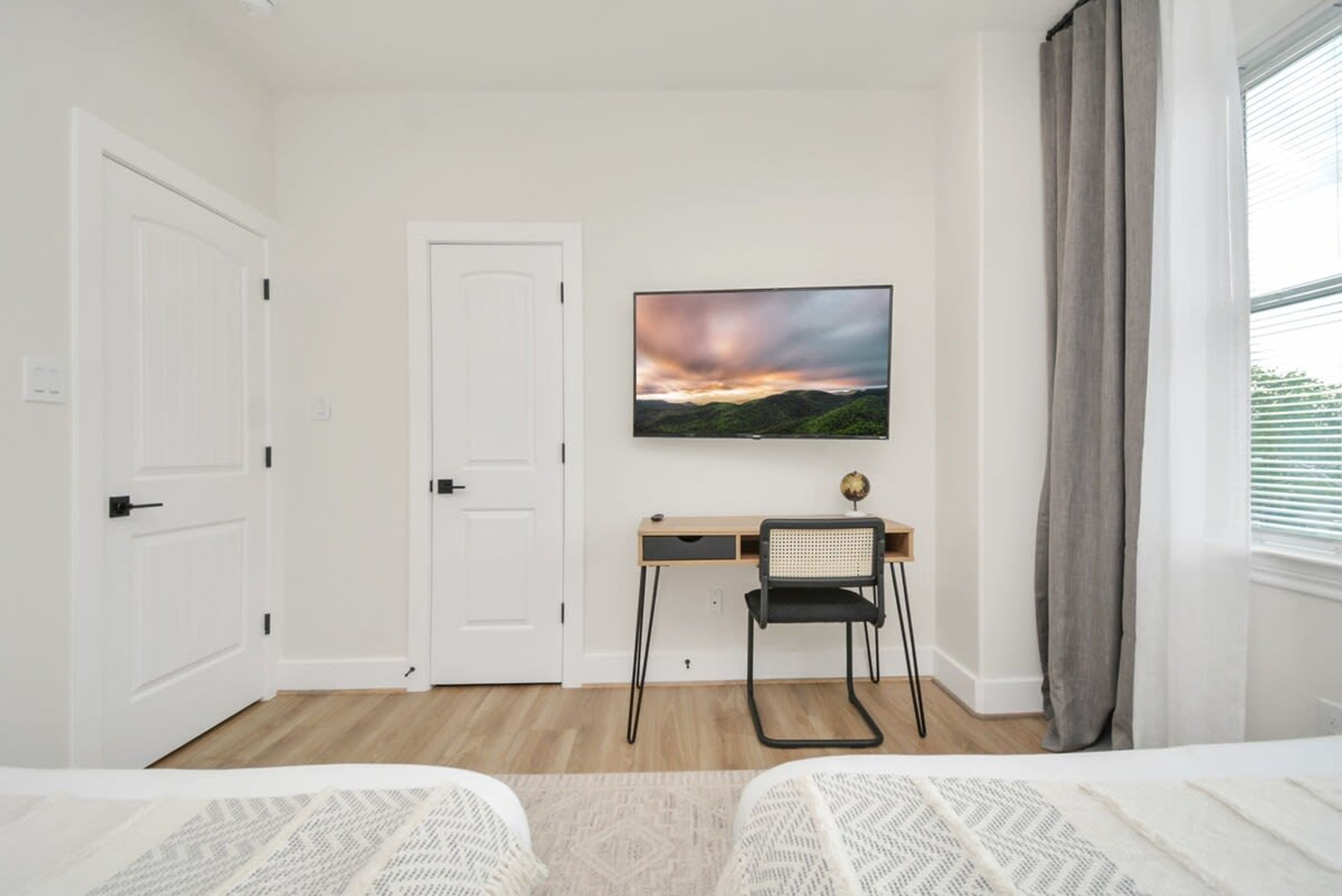 A bedroom with two beds, a desk with a chair, a painting on the wall, and a window with curtains.