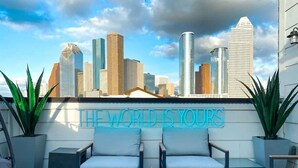 An outdoor seating area with two white cushioned chairs, a fire feature, and a neon sign stating "THE WORLD IS YOURS" against a city skyline backdrop. - Urban Jungle Retreat in the HTX Skyline (Houston)