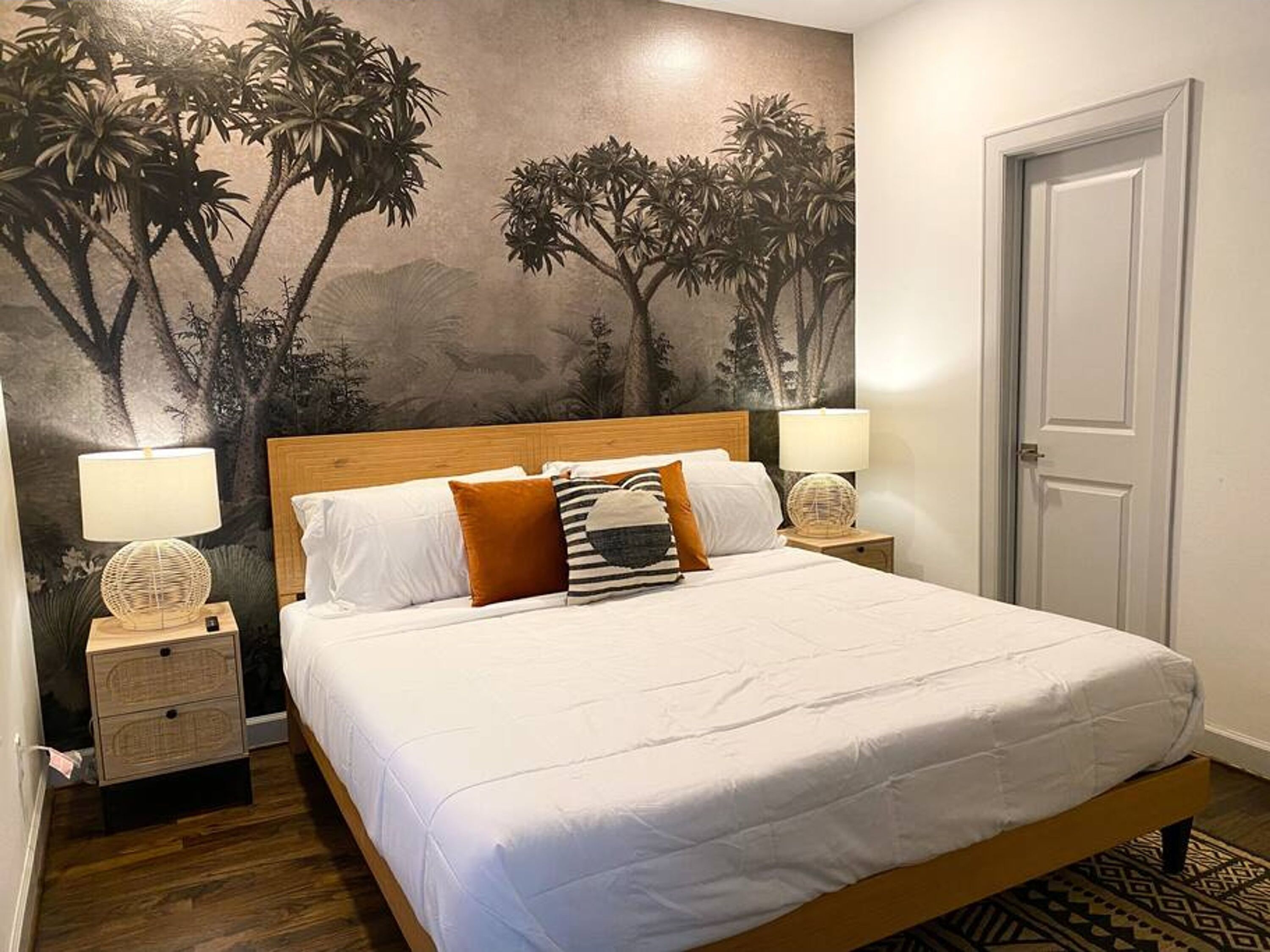 Una cama bien hecha con ropa de cama blanca y marrón, dos lámparas de noche y un mural de árboles en la pared.