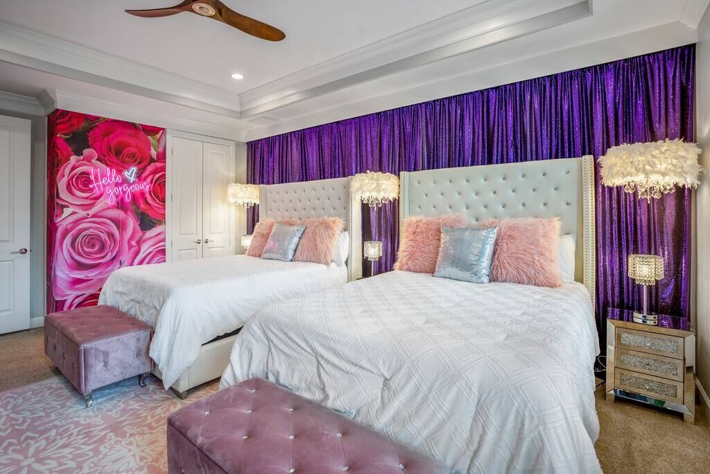 A bedroom with two beds, a pink ottoman, and a purple curtain backdrop.