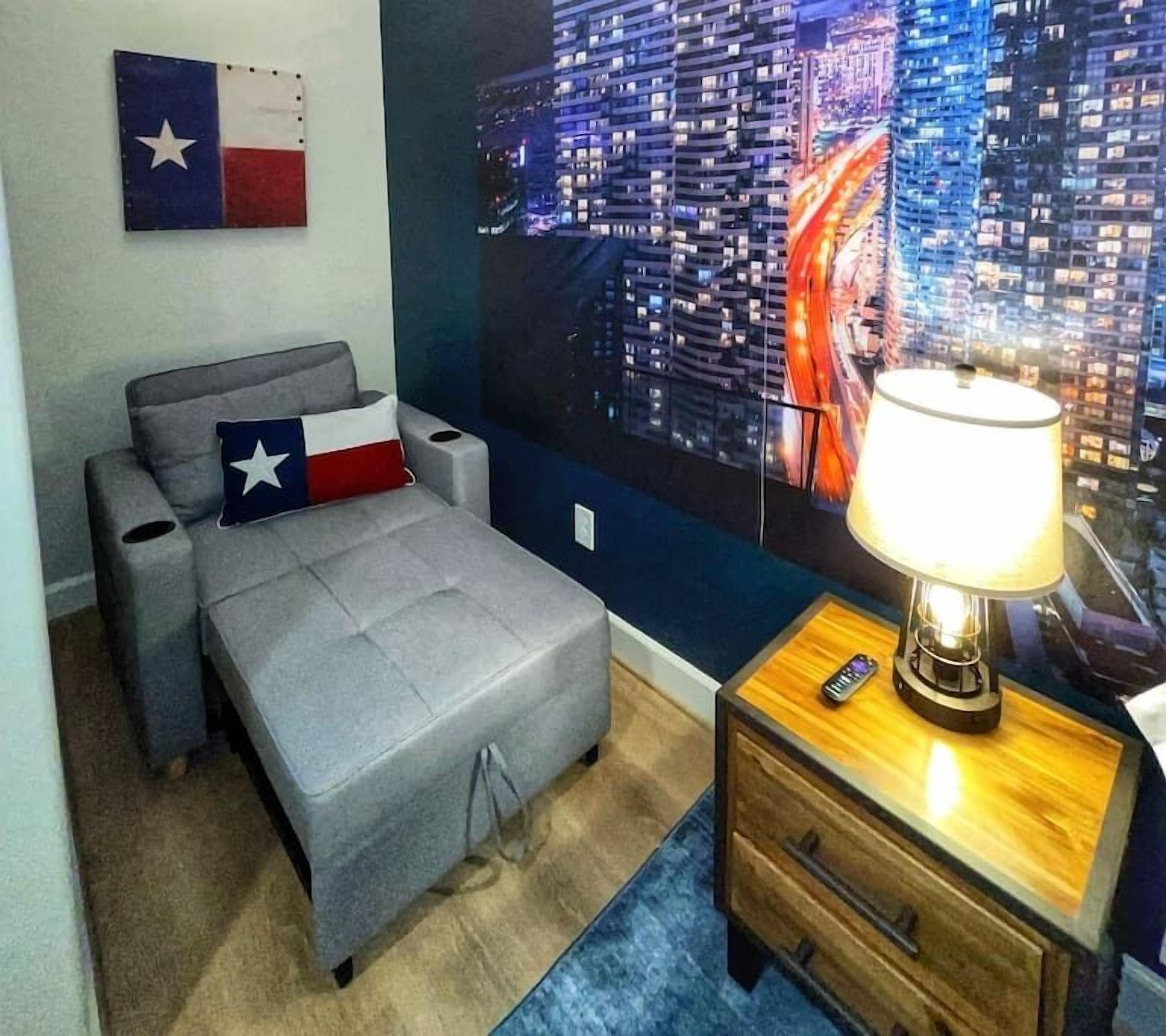 Een slaapkamer met een bed, nachtkastje, lamp, een Texas vlag en een stadsgezicht muurschildering.