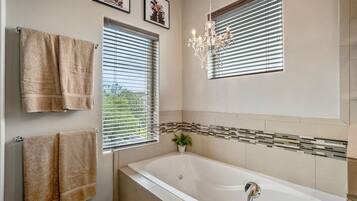 Un baño moderno con una bañera grande, dos toalleros, una araña y una ventana con persianas.