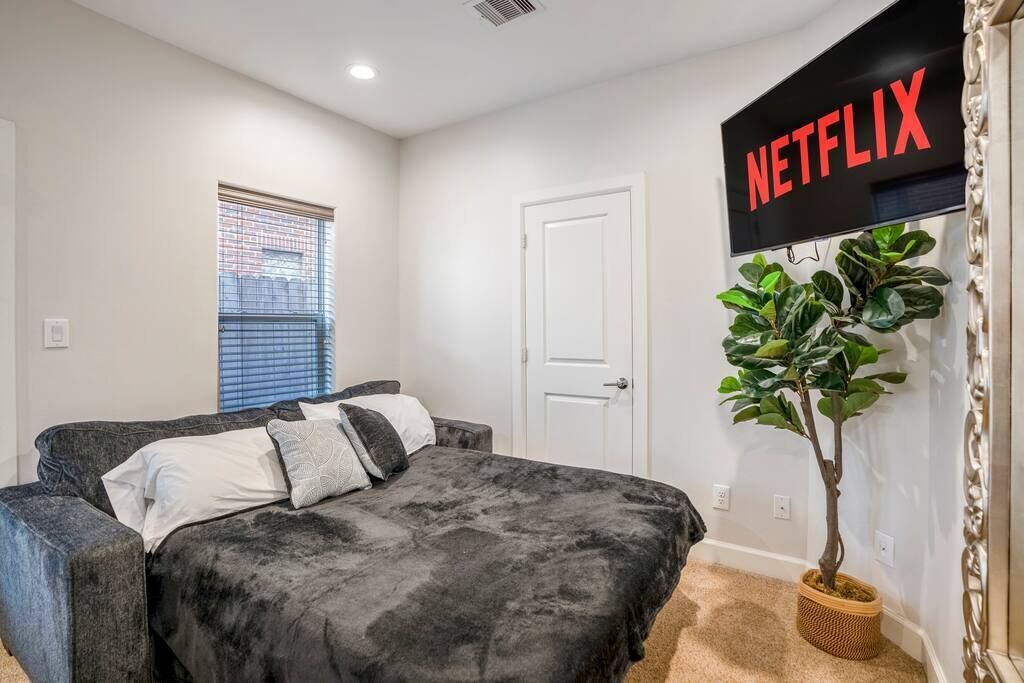 A bedroom with a bed, a wall-mounted TV, a door, and a window with blinds.