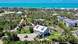 An aerial view of a coastal resort with multiple buildings, including a prominent white house with a blue roof, surrounded by lush greenery and palm trees.