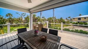 A covered outdoor area with a wooden table and chairs, a ceiling fan, and a view of palm trees and houses.