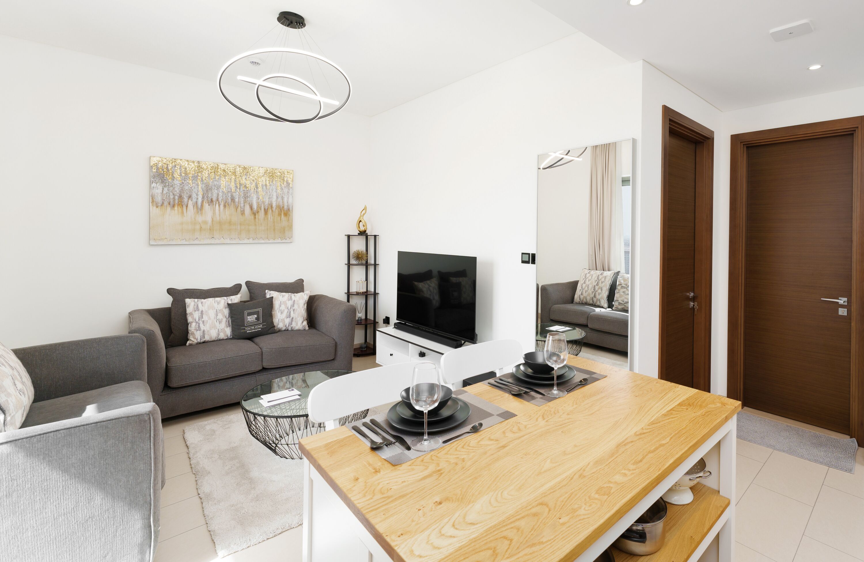 A modern living room with a grey sofa, a wooden dining table set for two, a flat-screen TV, and a large abstract painting on the wall.