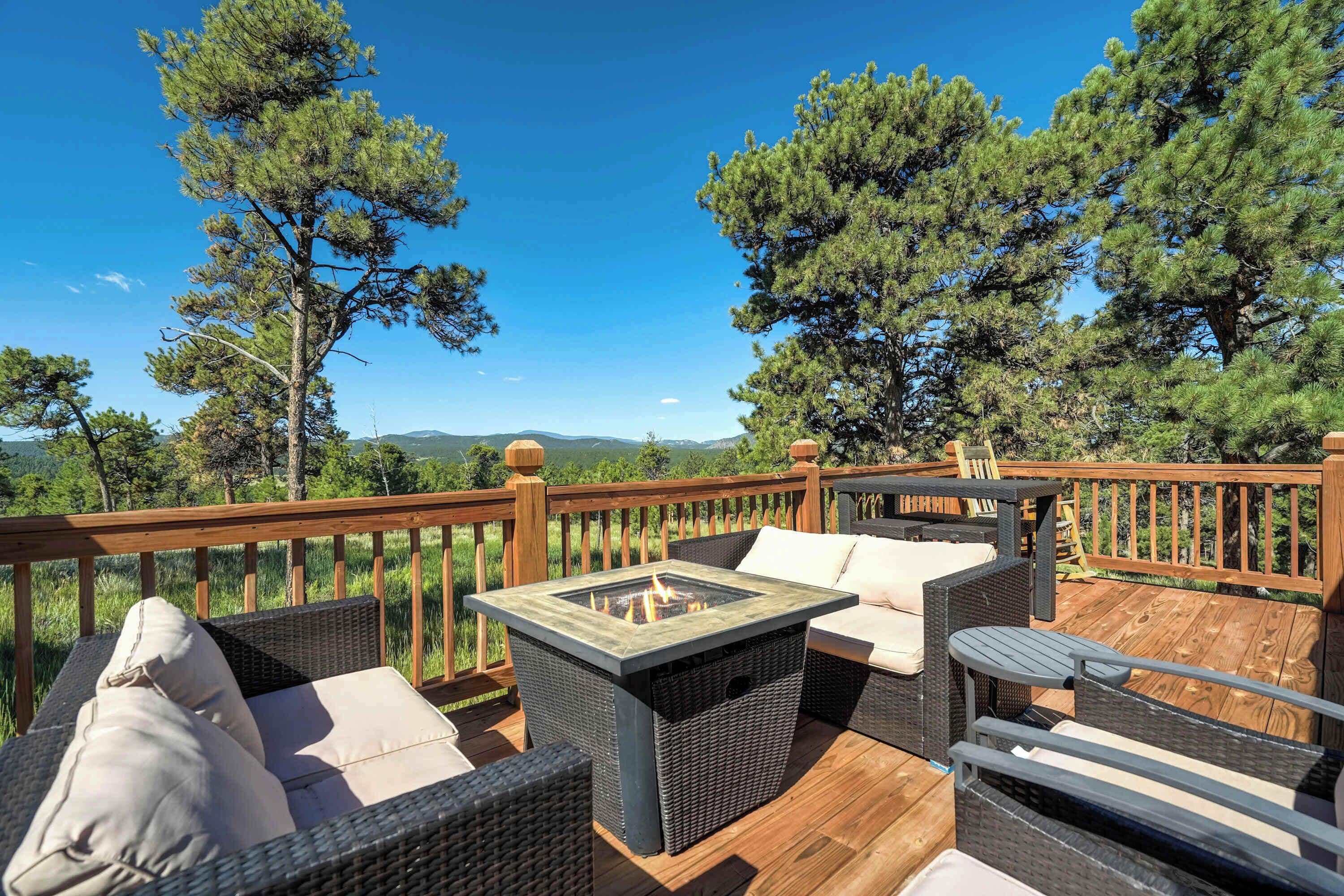 Una terraza al aire libre con mobiliario de mimbre, una mesa con superficie de vidrio y vistas a pinos y montañas lejanas.