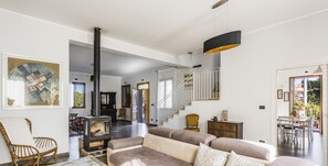 A spacious living area with a large beige sofa, a wooden coffee table, and a fireplace. There is a staircase leading to an upper level, and a dining area is visible in the background.