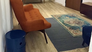 Un salon avec un plancher en bois, un fauteuil marron, une poubelle bleue, un tapis à motifs, un meuble en bois et une porte.