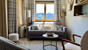 Un salon chaleureux avec un canapé, une petite table ronde et un fauteuil. Une fenêtre, ornée de rideaux, offre une vue sur les montagnes.