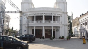 A white building with a balcony and columns, surrounded by parked cars and potted plants.