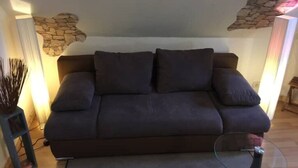 A dark-colored sofa with two cushions, a glass coffee table, and a stone wall decoration. - 2 Zimmer Wohnung/zentral/bayernähe! (Leverkusen)