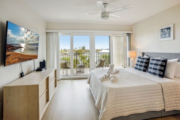 Quarto de hotel com uma TV grande, uma cama com cobertura acolchoada, um ventilador de teto, uma varanda com cadeiras e vista para a praia.