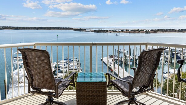 A balcony with wicker chairs and a small table, overlooking a marina with boats and a distant shoreline.