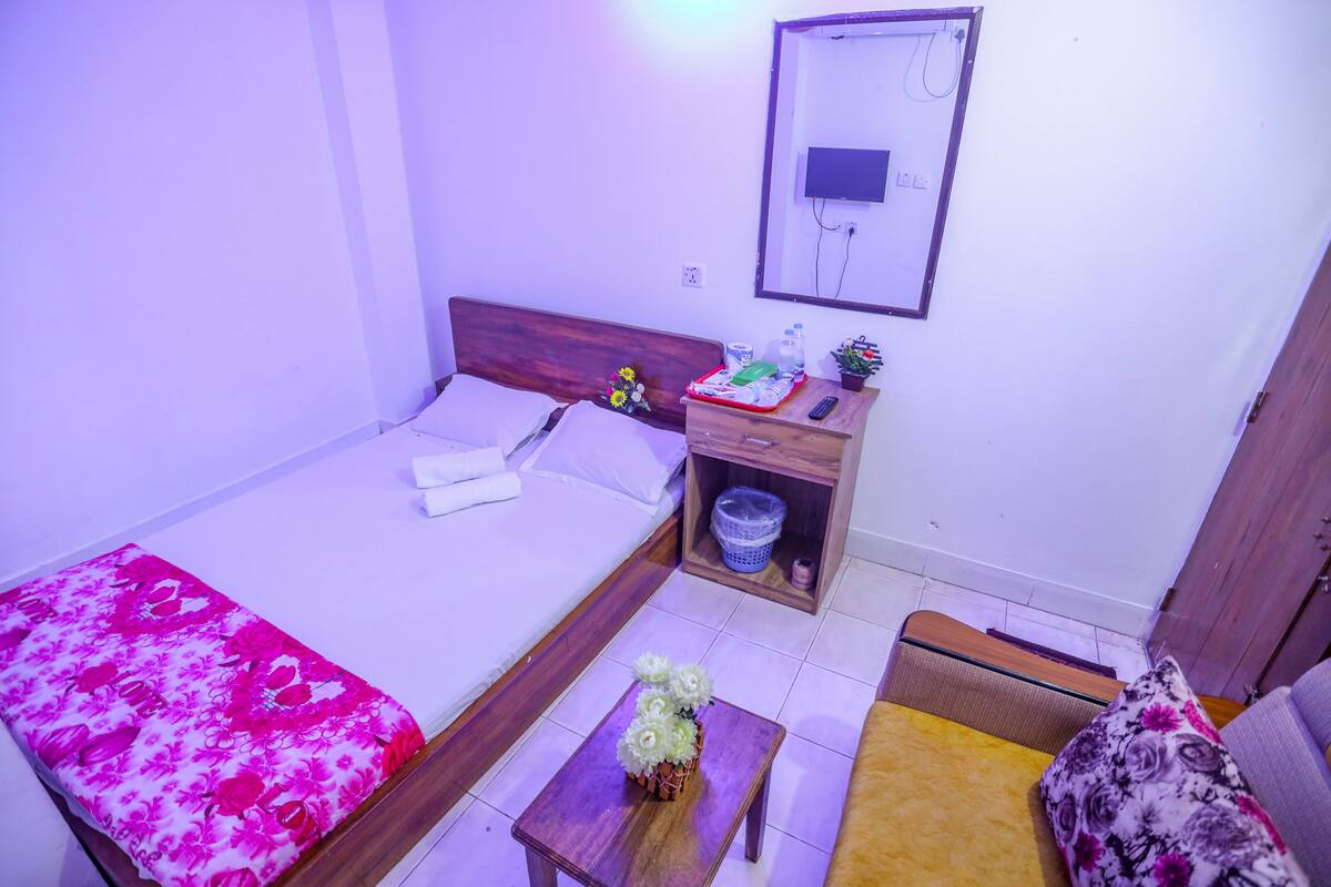 A hotel room with a bed, a small table with a vase of flowers, a wooden nightstand, a sofa, and a wall-mounted TV.