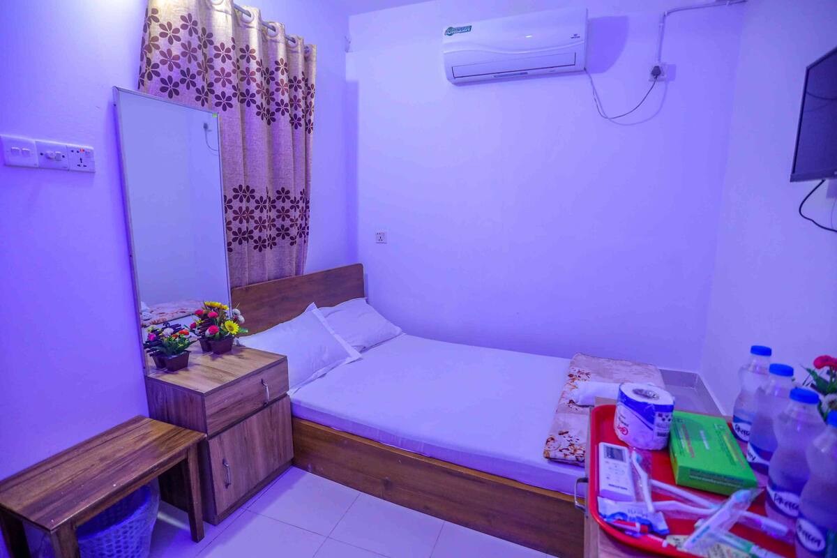 A bedroom with a bed, a wooden nightstand, a mirror, a window with curtains, an air conditioning unit, and a desk with various items.