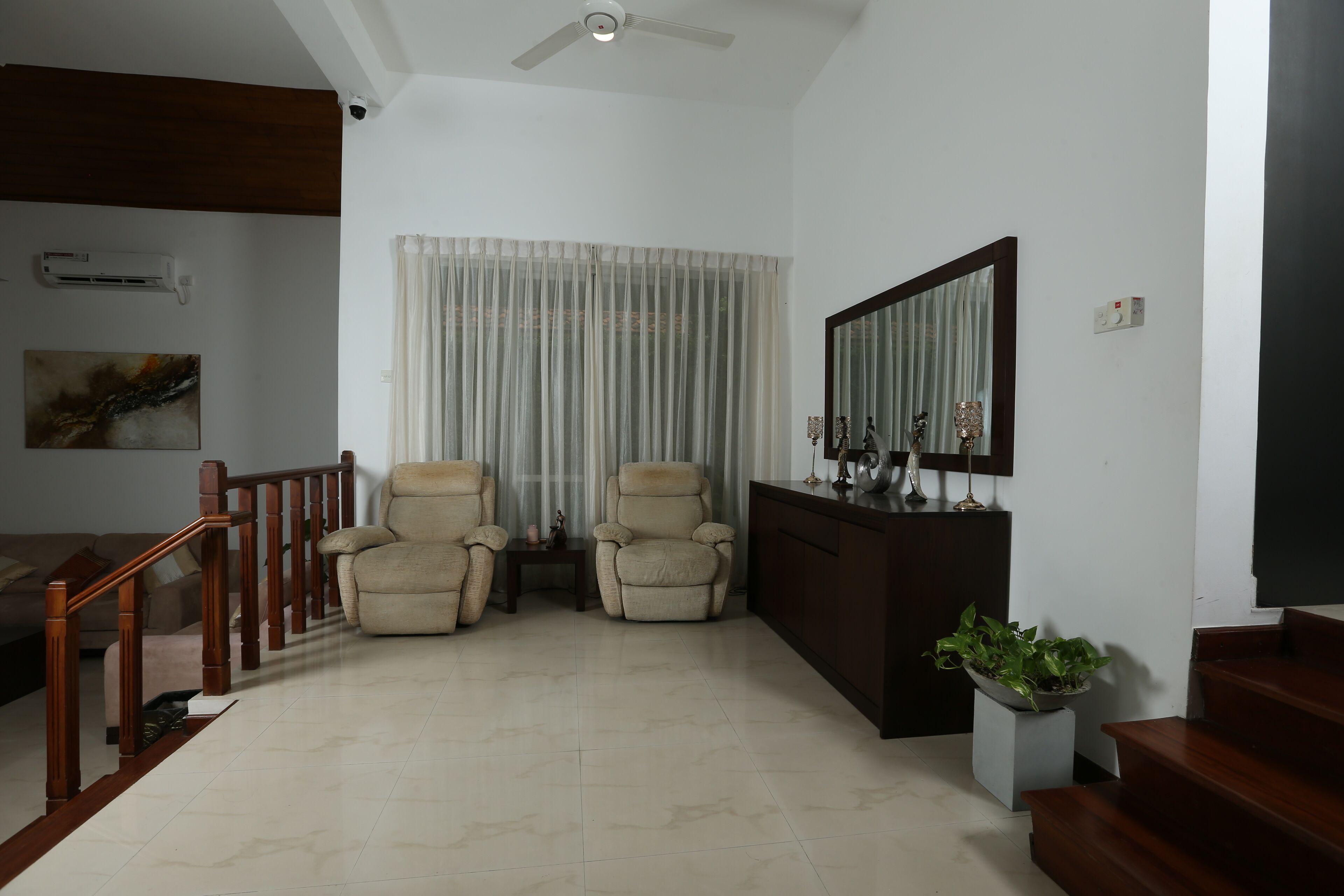 A living room with a wooden staircase, two beige armchairs, a small table, a potted plant, and a large mirror on the wall.