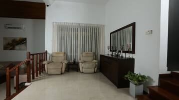 A living room with a wooden staircase, two beige armchairs, a small table, a potted plant, and a large mirror on the wall.