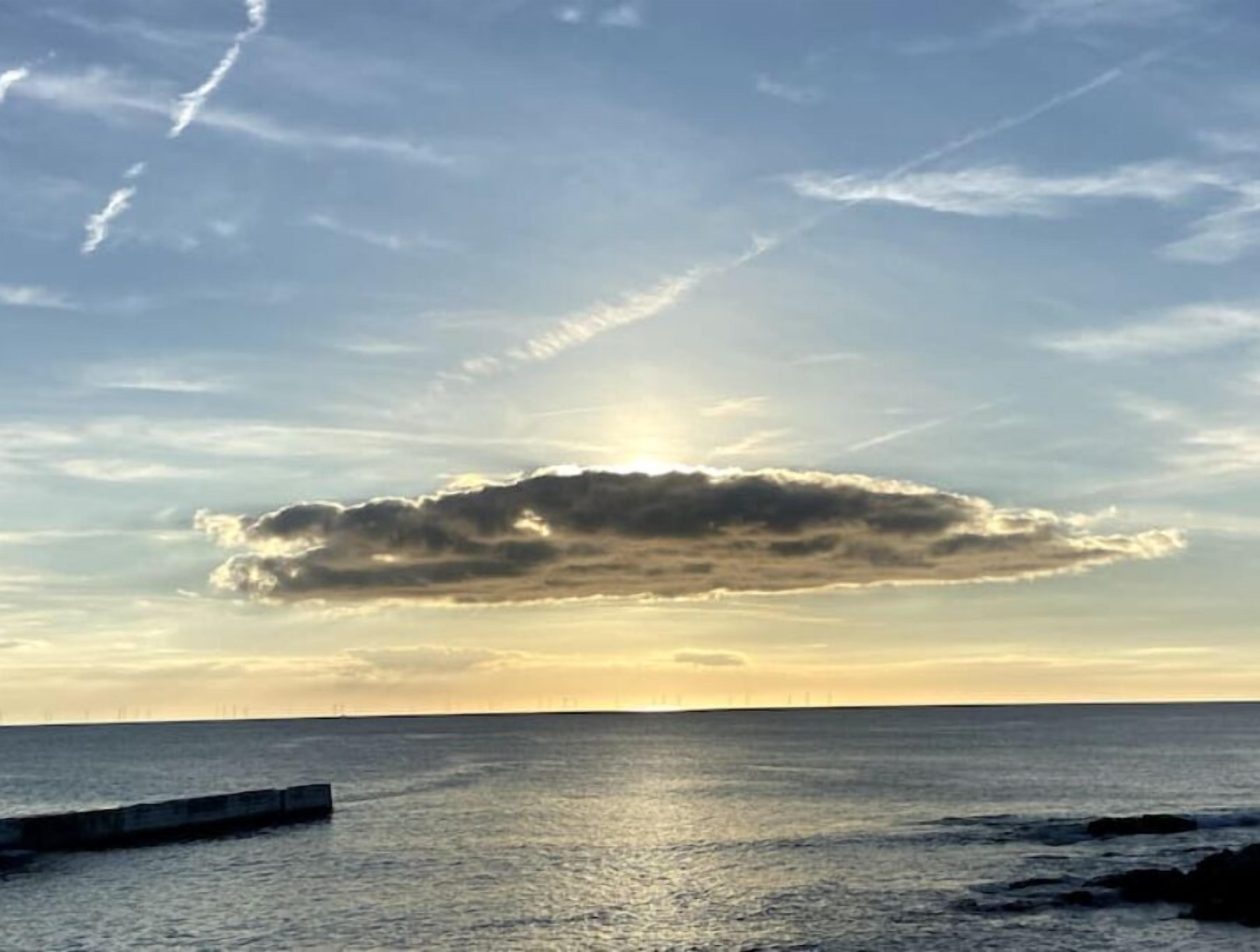 Paesaggio costiero con mare calmo, molo in cemento e tramonto con nuvole.