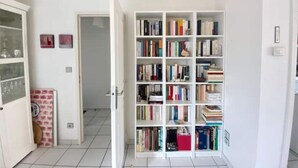 A room with a white tiled floor, a white bookshelf filled with books, a white door, and a white wall with a small picture. - Maison Avec Plages et Commerces à Pieds (Batz-sur-Mer)